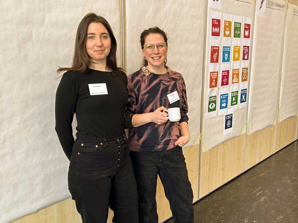 Två personer står mot en vägg med globala målen.