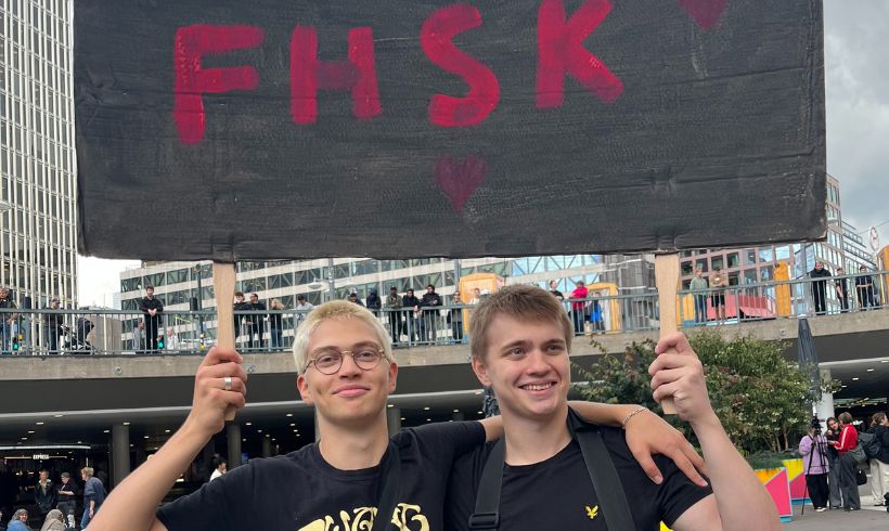 Två personer står och håller ett plakat som det står FHSK på.